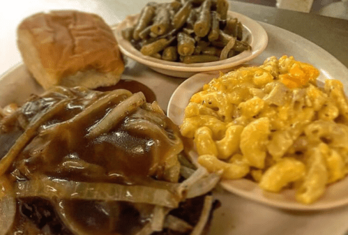 A plate featuring a sandwich with onions and gravy, macaroni and cheese, and green beans, alongside a roll.