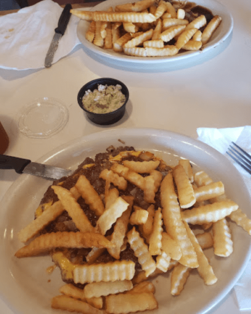 Two plates of crinkle-cut fries, one topped with ground beef and cheese, and a small bowl of green sauce on the side.