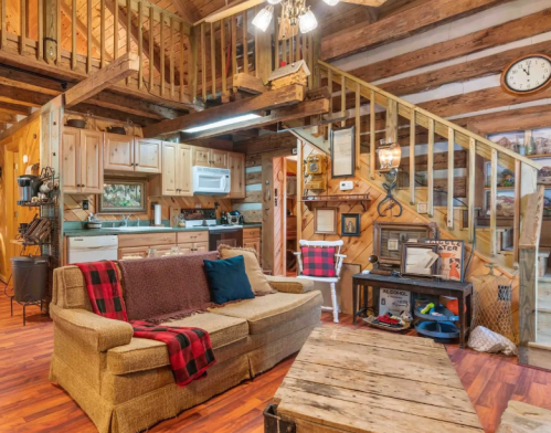 Cozy cabin interior featuring a sofa, wooden decor, kitchen area, and a staircase leading to a loft. Warm, rustic ambiance.