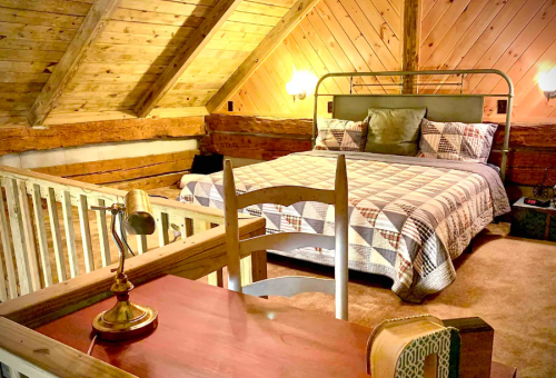 Cozy attic bedroom with a wooden ceiling, a queen bed, and a desk with a lamp, featuring rustic decor.
