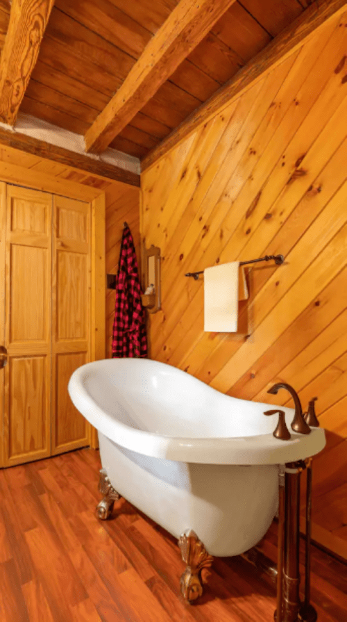 A cozy bathroom featuring a vintage clawfoot tub, wooden walls, and a rustic decor with a plaid robe hanging nearby.