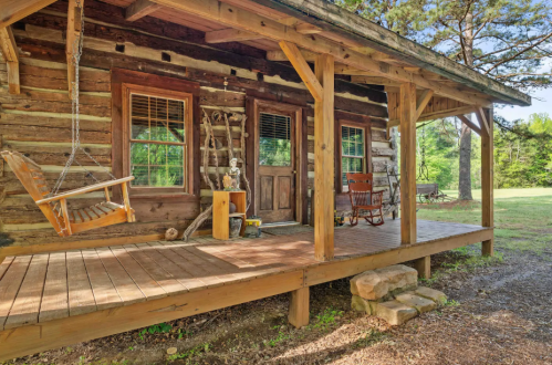 A rustic log cabin with a wooden porch, featuring a swing and rocking chairs, surrounded by greenery.