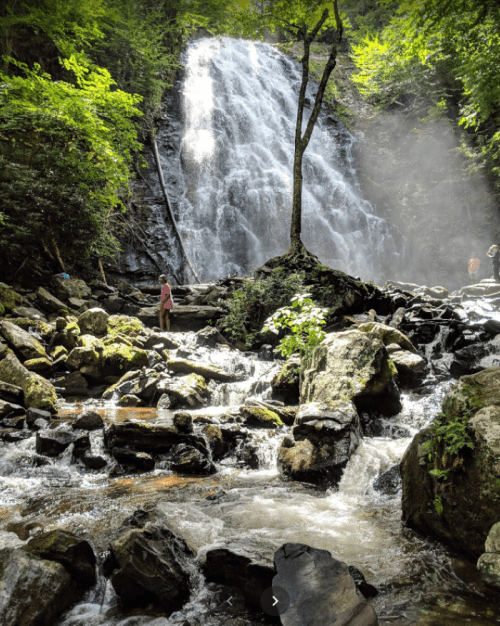 A person stands near a waterfall surrounded by lush greenery and rocky terrain, with mist rising from the water.