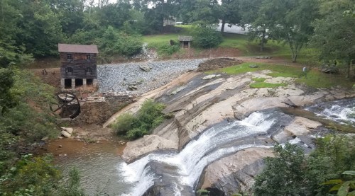 A scenic view of a waterfall and an old wooden mill surrounded by greenery and rocky terrain.
