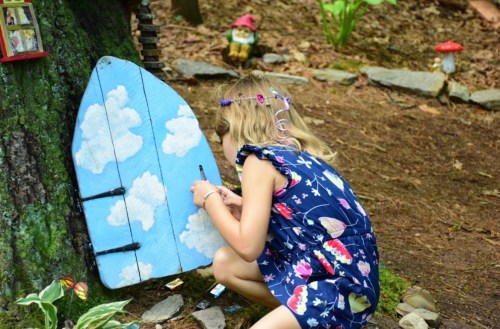 A child in a blue dress paints a whimsical door on a tree, surrounded by nature and small decorations.