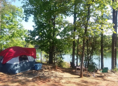 A red and gray tent by a serene lake, surrounded by trees and camping chairs on a sunny day.