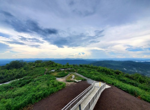 A scenic view from a mountain overlook, featuring lush greenery and a cloudy sky in the distance.