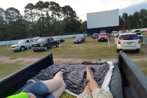 A view from the back of a truck at a drive-in movie, with people relaxing and cars parked in front of a large screen.