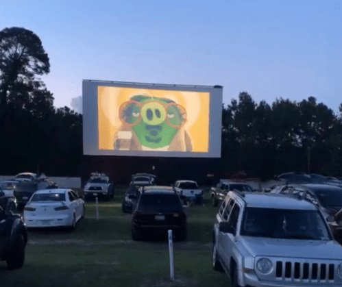 A drive-in movie screen shows an animated character with glasses, surrounded by parked cars in a grassy area.