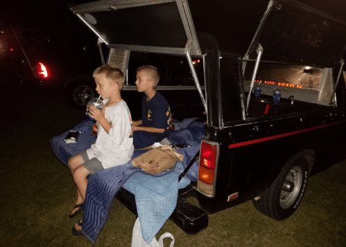 Two boys sit on the back of a truck at night, enjoying snacks and drinks, with a blanket draped over the tailgate.
