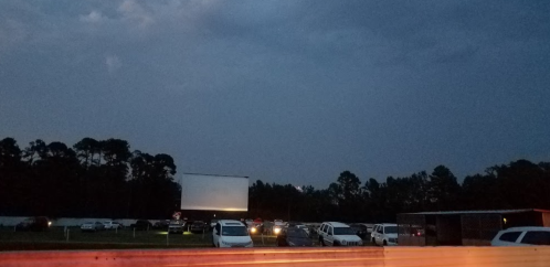 A drive-in movie theater at dusk, with cars parked and a large blank screen in the background under a cloudy sky.