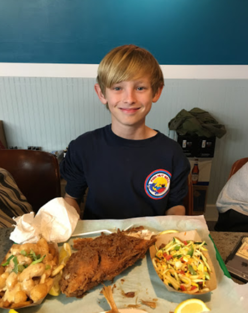 A smiling boy holds a tray with fried fish, shrimp, and a side of salad in a casual dining setting.