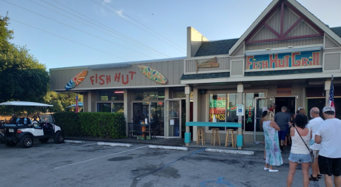 A busy seafood restaurant with a sign reading "Fish Hut" and a line of customers outside. Golf cart parked nearby.