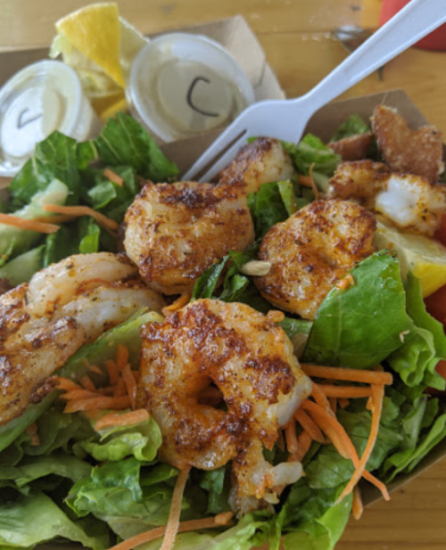 A fresh salad topped with seasoned shrimp, carrots, and lettuce, served with two small containers of dressing and lemon wedges.