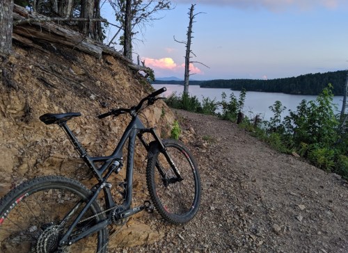 A mountain bike rests on a dirt path overlooking a calm lake and distant hills at sunset.
