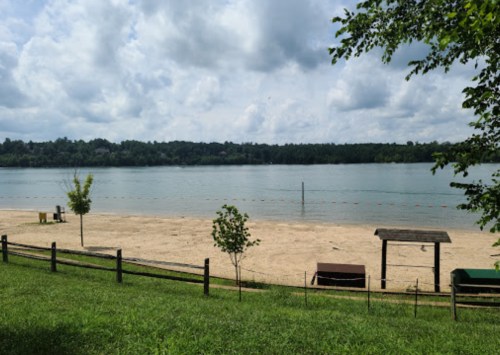 A serene beach scene with sandy shore, calm water, and trees under a cloudy sky. A picnic area is visible nearby.