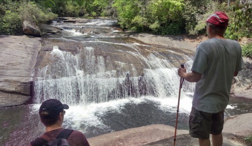Two hikers stand by a waterfall, surrounded by lush greenery, enjoying the serene natural landscape.