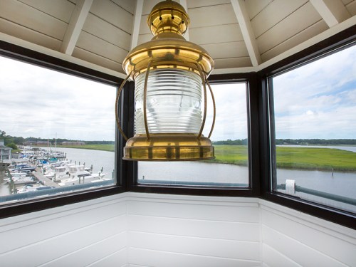 A brass lantern hangs inside a lighthouse, with a view of a marina and river through large windows.