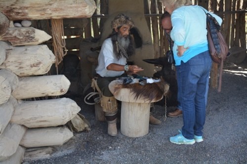 A person in traditional attire demonstrates crafting tools to a visitor at a historical site.