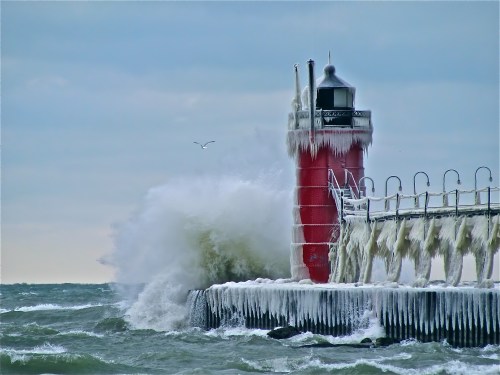 A red lighthouse covered in ice, with waves crashing against the pier under a cloudy sky.