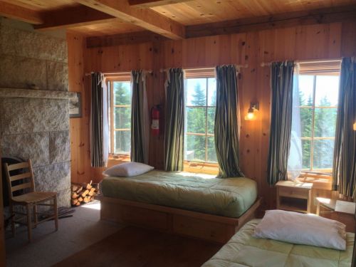 Cozy wooden bedroom with two beds, large windows, and a chair, surrounded by trees outside.
