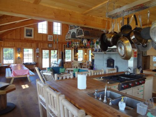 Cozy wooden kitchen and dining area with a large table, hanging pots, and bright windows. Warm, inviting atmosphere.