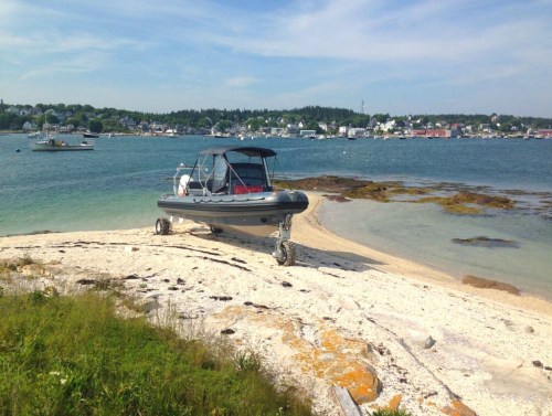 A small boat on a sandy shore with calm waters and a coastal town in the background under a clear blue sky.
