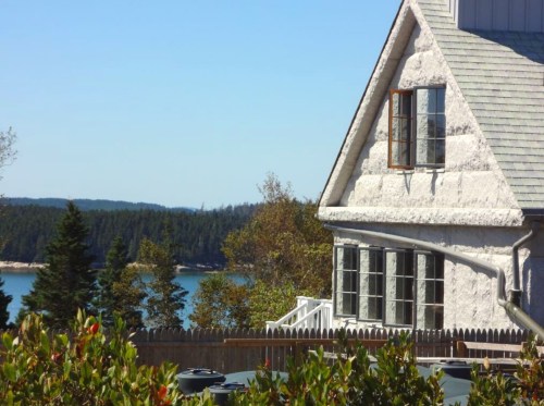 A coastal house with large windows overlooks a serene body of water, surrounded by trees and a wooden fence.
