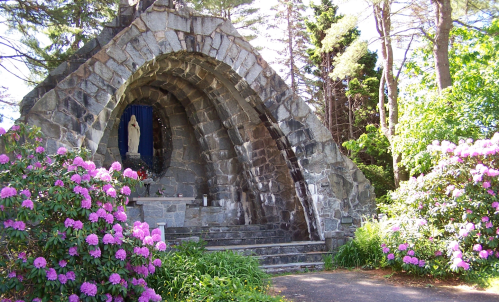 A stone archway with a statue of Mary, surrounded by vibrant pink flowers and lush greenery.