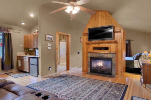 Cozy living space with a fireplace, kitchen area, and wooden accents, featuring a rug and ceiling fan.