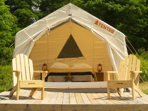 A cozy glamping tent with a bed, two wooden chairs, and lanterns, set in a natural outdoor environment.