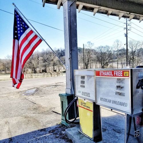 A gas station pump with a U.S. flag in the background, displaying "Ethanol Free" and "Regular Unleaded" labels.