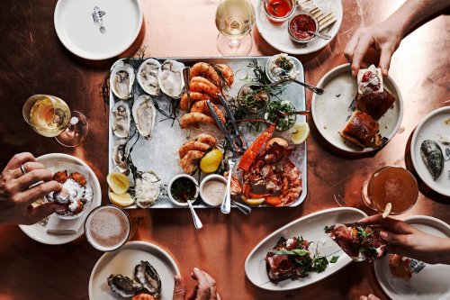 A table spread with seafood, including oysters, shrimp, and lobster, surrounded by drinks and hands reaching for food.