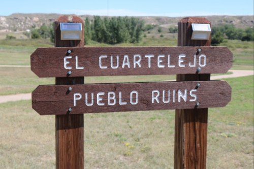 Wooden signpost with two directional signs: "El Cuartelejo" and "Pueblo Ruins," set in a grassy landscape.
