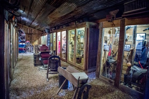 Interior of a rustic museum displaying vintage horse-drawn carriages, saddles, and historical photographs in glass cases.