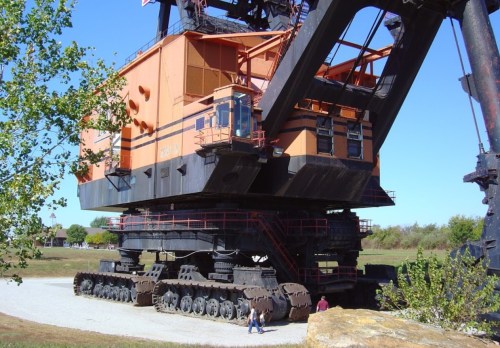 A large orange industrial excavator on tracks, with people standing nearby for scale against a clear blue sky.