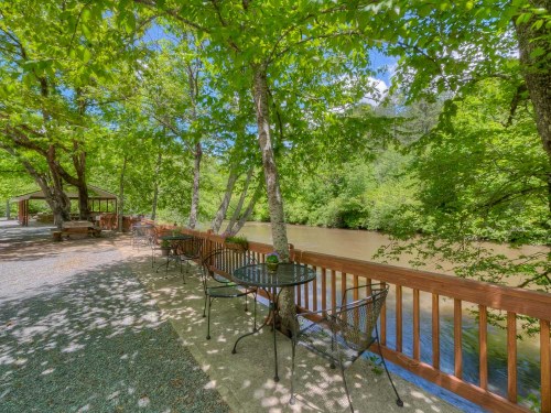 A serene riverside scene with tables under trees, surrounded by lush greenery and a clear blue sky.