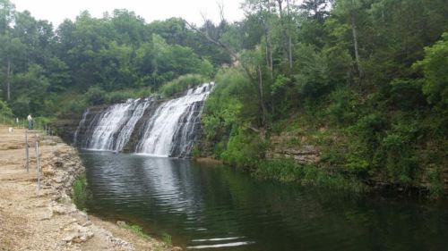 A serene waterfall cascading into a calm pool, surrounded by lush greenery and rocky terrain.