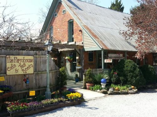 A charming brick farmhouse café with a sign, colorful flowers, and a welcoming entrance surrounded by greenery.