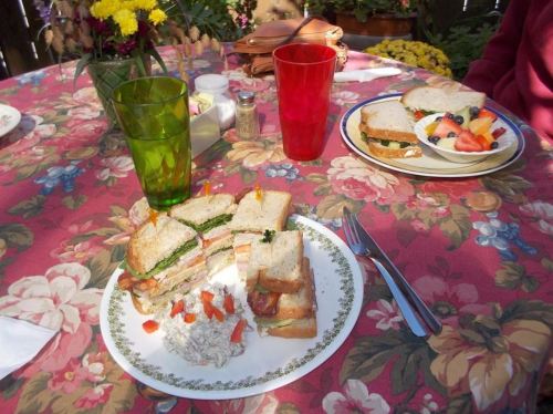 A table set with colorful drinks, sandwiches, and a fruit plate, surrounded by flowers and a patterned tablecloth.