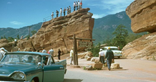 A vintage scene with people on a rock formation, cars parked nearby, and a sign for "Balanced Rock" in a scenic landscape.