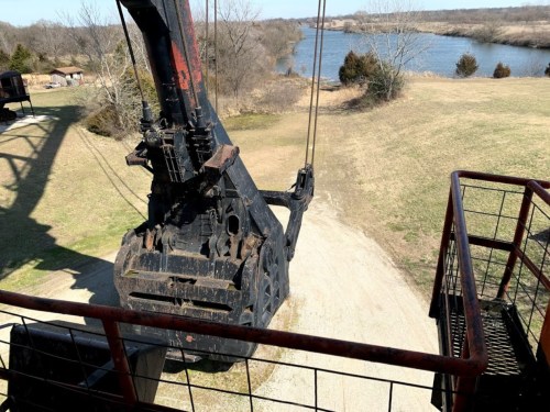A large excavator bucket overlooks a grassy area and a river in the background on a clear day.