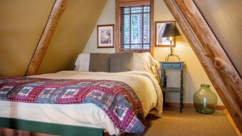 Cozy attic bedroom with a bed, plaid blanket, wooden beams, and a bedside table with a lamp. Natural light from a window.