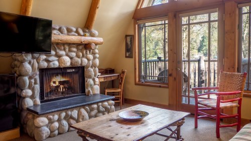 Cozy cabin interior with a stone fireplace, wooden furniture, and large windows overlooking a forested view.
