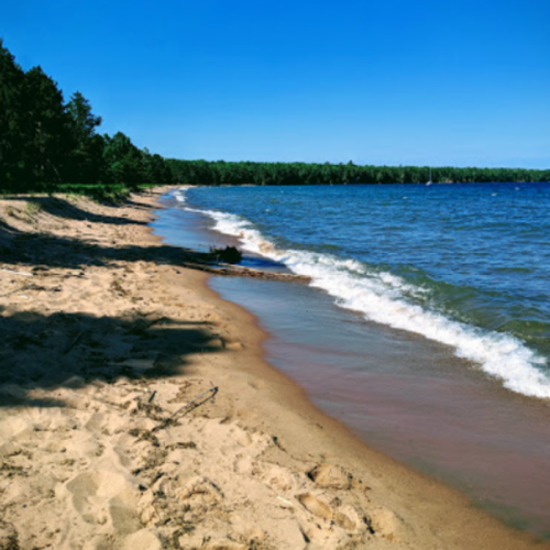 A sandy beach curves along a calm lake, surrounded by green trees under a clear blue sky.