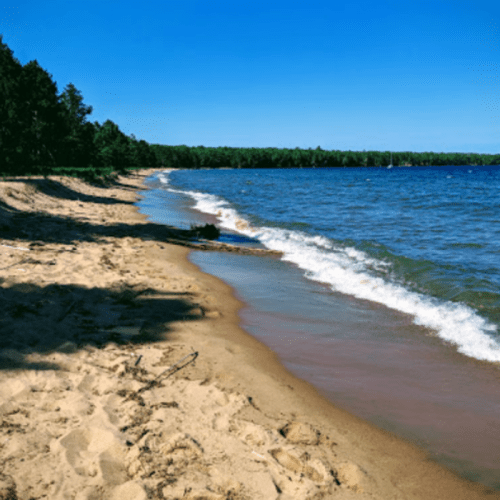 A sandy beach curves along a calm lake, surrounded by green trees under a clear blue sky.