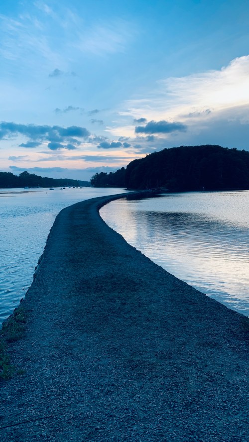 A winding gravel path along a calm river at dusk, surrounded by trees and a cloudy sky.