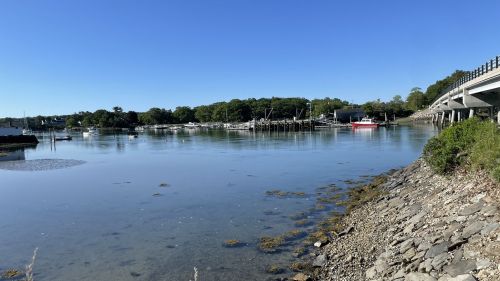 A calm river scene with boats, greenery, and a clear blue sky, featuring a rocky shoreline and a bridge in the background.