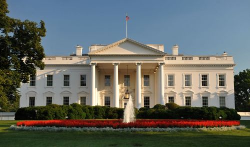 The White House, a large white building with columns, surrounded by green bushes and red flower beds, under a clear sky.