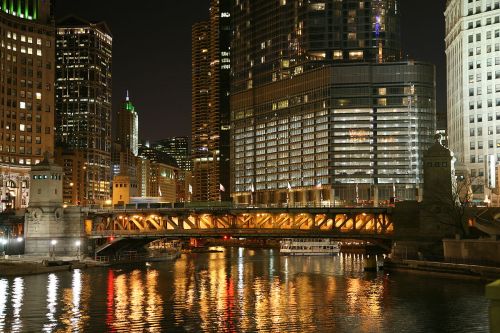 A city skyline at night, featuring a lit bridge over a river, with reflections of buildings in the water.